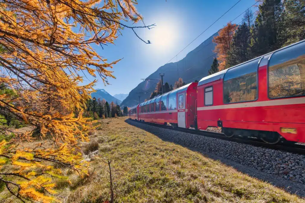 A red train travels through a scenic landscape with autumn trees and mountains under a bright sun.