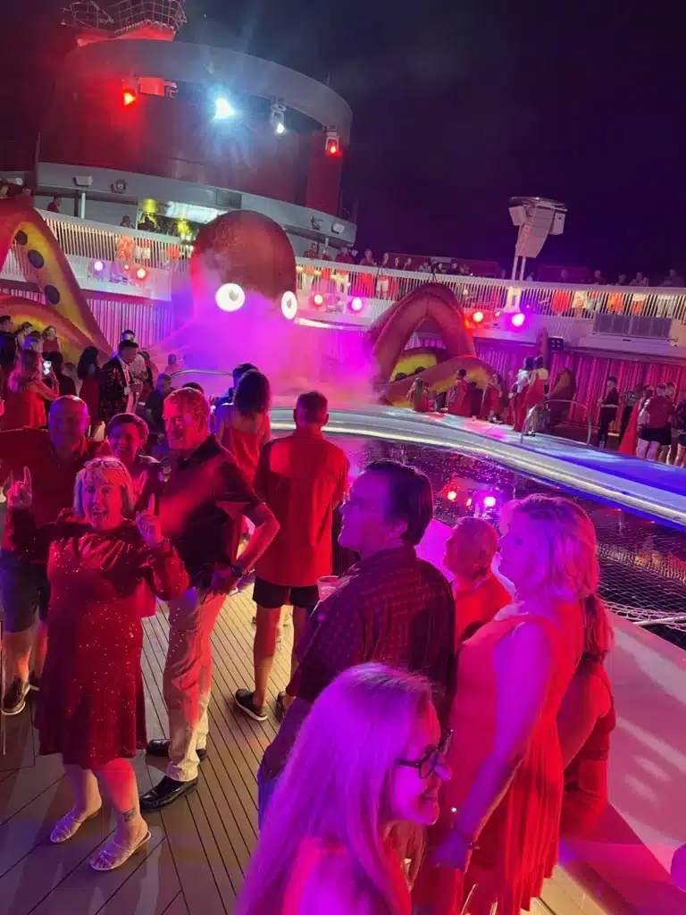 People in red outfits are enjoying a lively Virgin Voyages cruise party on the ship's deck at night, surrounded by colorful lights and an eye-catching octopus decoration in the background.