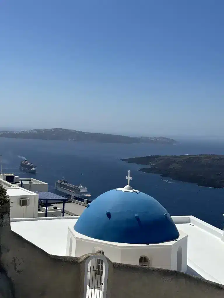 Perched gracefully above the Aegean Sea, the blue-domed building offers a breathtaking view of cruise ships, including Virgin Voyages vessels, and scattered islands under a clear blue sky.