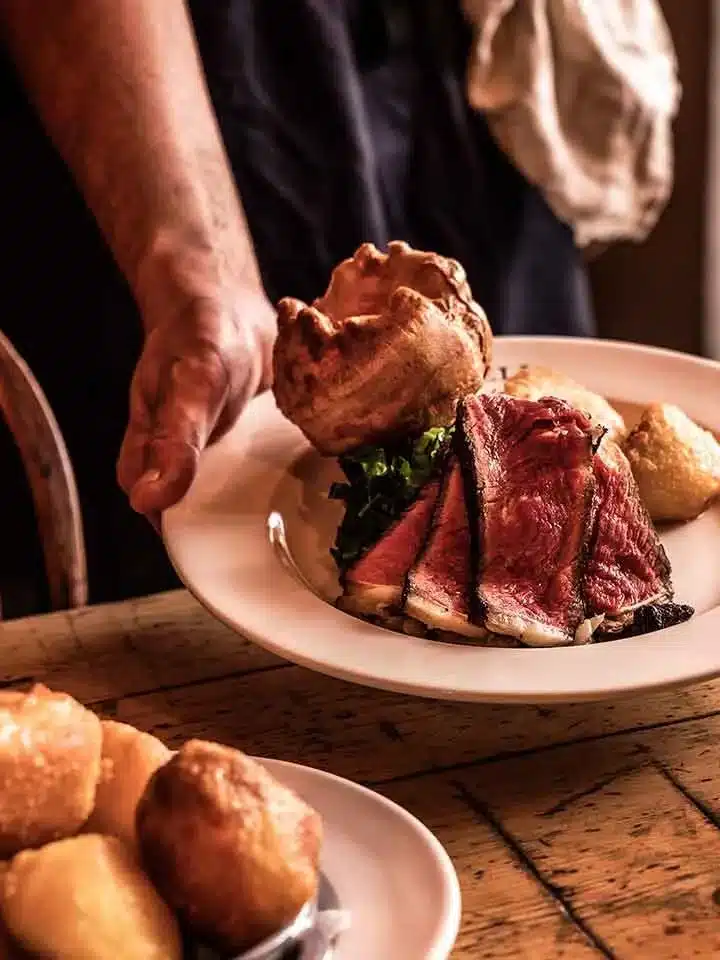 A person serves a plate with sliced beef, a Yorkshire pudding, and vegetables—reminiscent of the hearty meals found at the best Cotswold pubs. Meanwhile, another plate with small round pastries sits enticingly on the table.