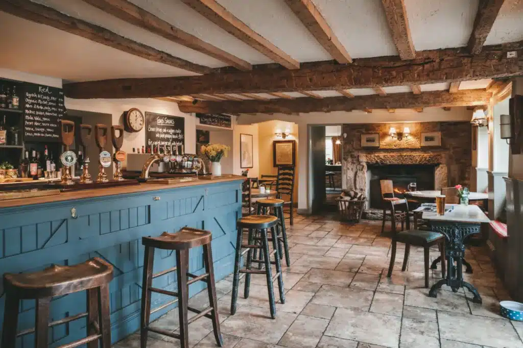 Cozy pub interior with wooden beams, a blue bar counter with stools, rustic tables, and a welcoming fireplace in the background. This charming setting is reminiscent of the best Cotswold pubs.