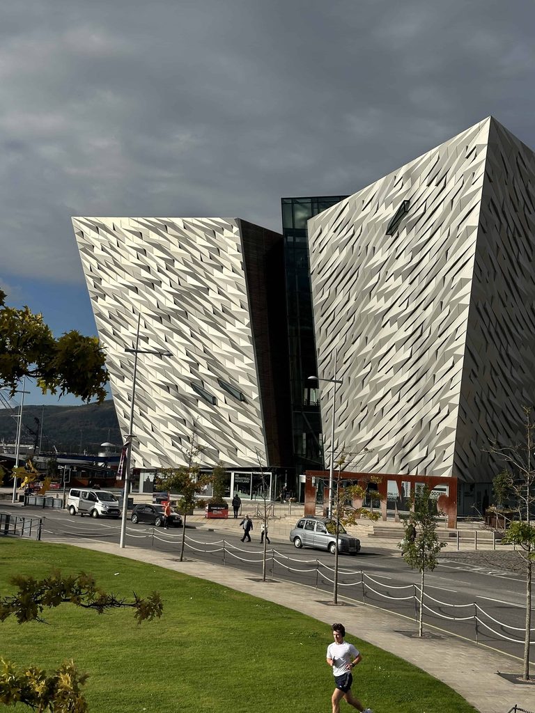 The Titanic Belfast museum, one of the best things to do in Belfast, features a geometric metallic exterior beside a street with parked cars and a pedestrian.