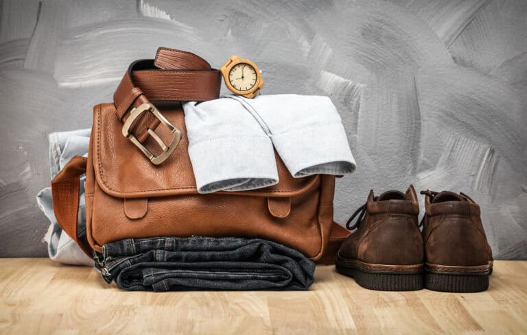 Leather bag with a belt and watch on top, resting on a folded blue shirt and dark jeans. Part of a men's packing list for Europe, the ensemble is completed by brown shoes placed beside the bag on a wooden surface against a gray background.