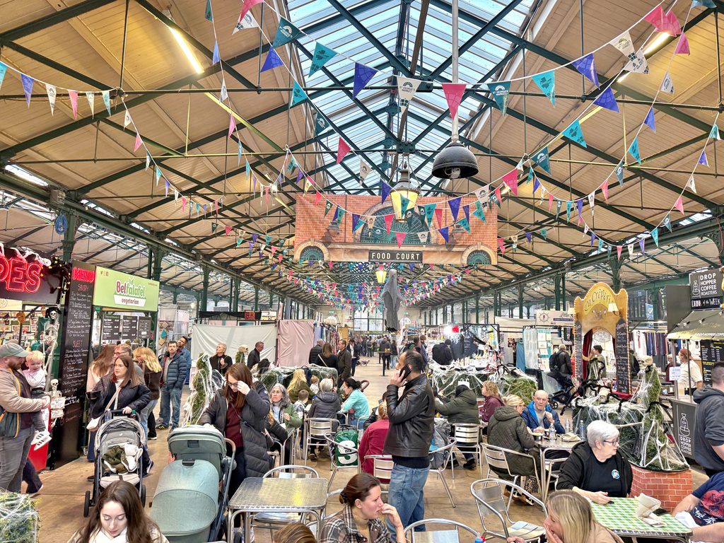Indoor food court with vendor stalls and dining tables under a high glass ceiling decorated with colorful triangular bunting, one of the best things to do in Belfast.