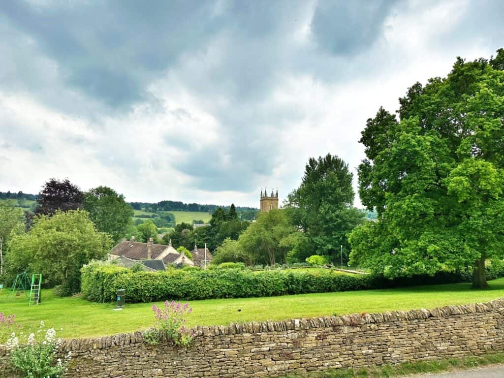 A village scene with a stone wall in the foreground, lush green trees, a church tower in the distance, and a cloudy sky above.