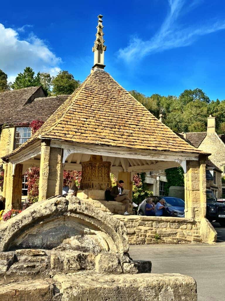 A stone gazebo with a tiled roof in a village square. People are sitting inside. Historic stone building in the background and trees under a blue sky.