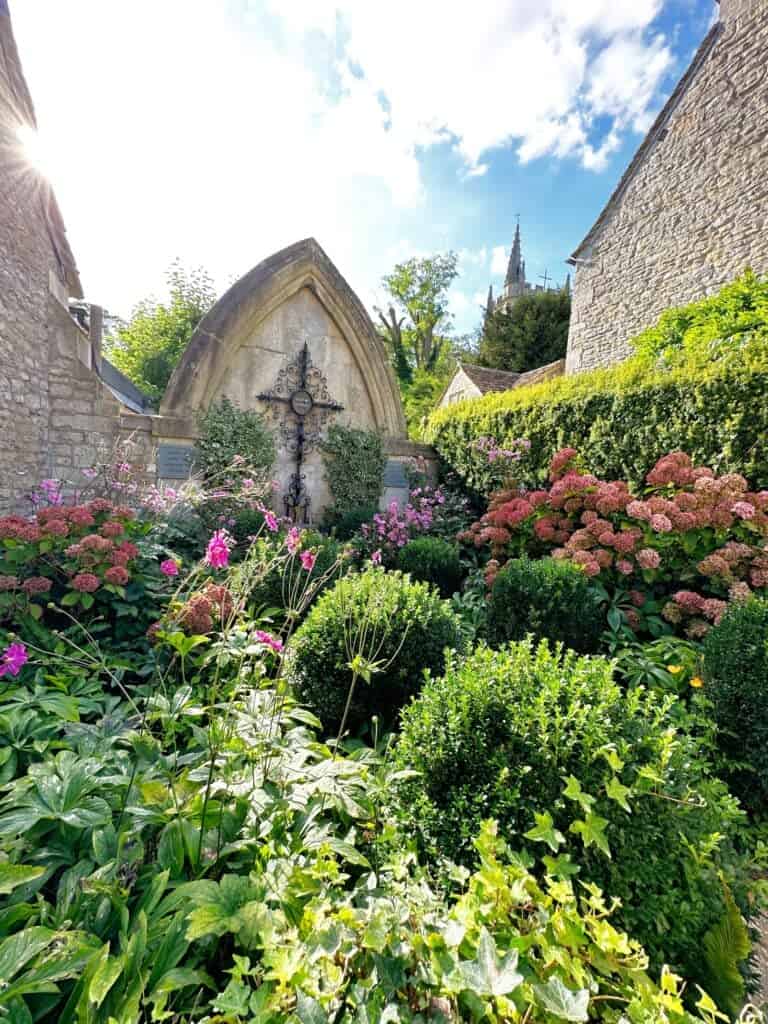 A garden with a variety of green plants and colorful flowers surrounds a stone structure with a cross. Stone buildings and a pointed tower are visible in the background under a blue sky.