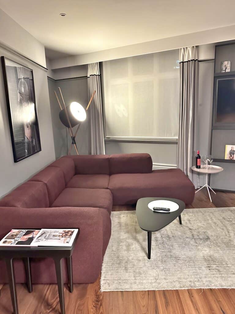Modern living room with a maroon sectional sofa, tripod floor lamp, two small tables, magazines, a wine bottle, and a remote on a rug over wooden flooring.