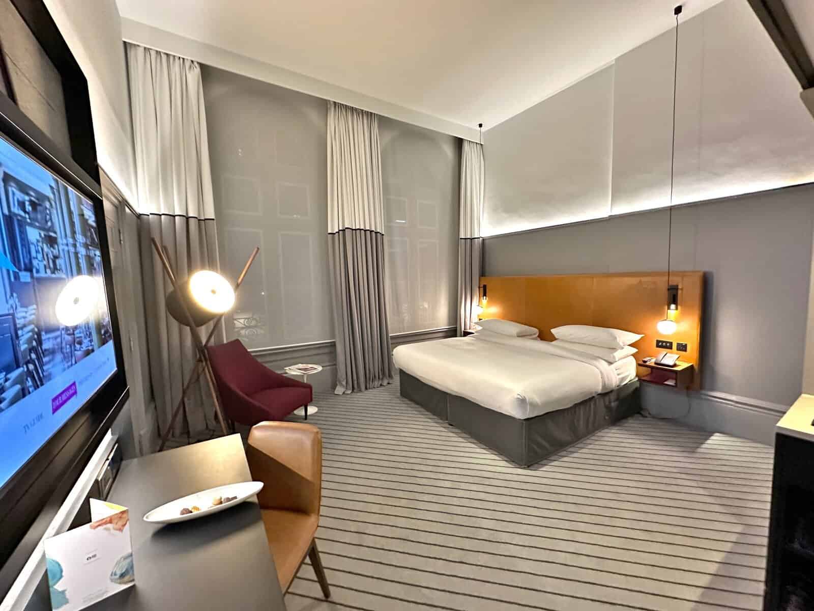 A king bed, television, desk, and lounge chair by the window in an Andaz London Liverpool Street hotel room.