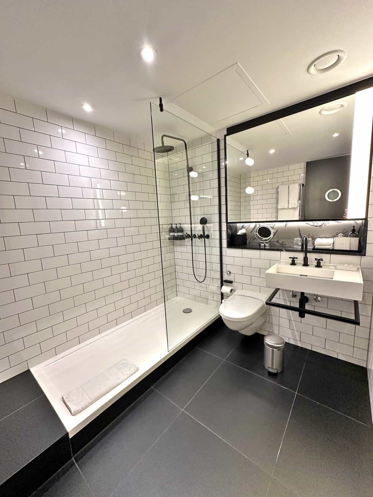 Modern bathroom at the Andaz London Liverpool Street featuring a glass-enclosed shower, wall-mounted toilet, and a white rectangular sink under a large mirror.