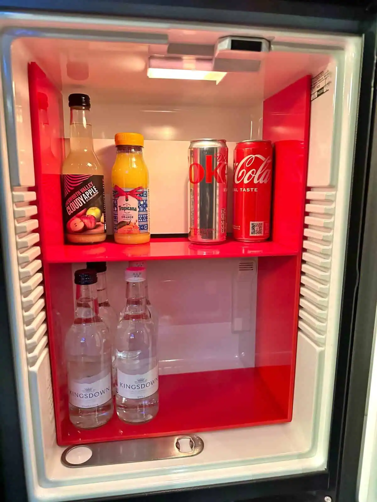 Open mini-fridge at the Andaz London Liverpool Street containing bottles of cloudy apple juice, orange juice, and sparkling water, along with cans of Diet Coke and Coca-Cola.