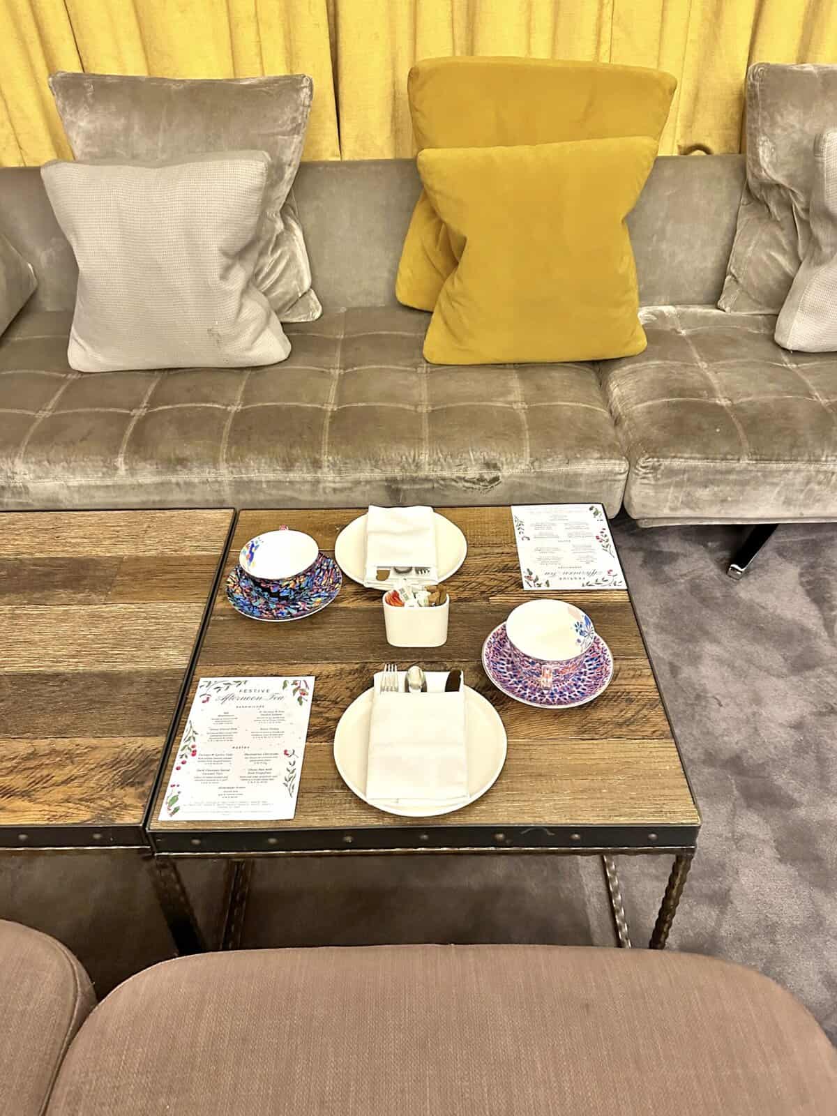A low wooden coffee table set for afternoon tea at the Andaz London Liverpool Street, positioned in front of a sofa with grey and yellow cushions.