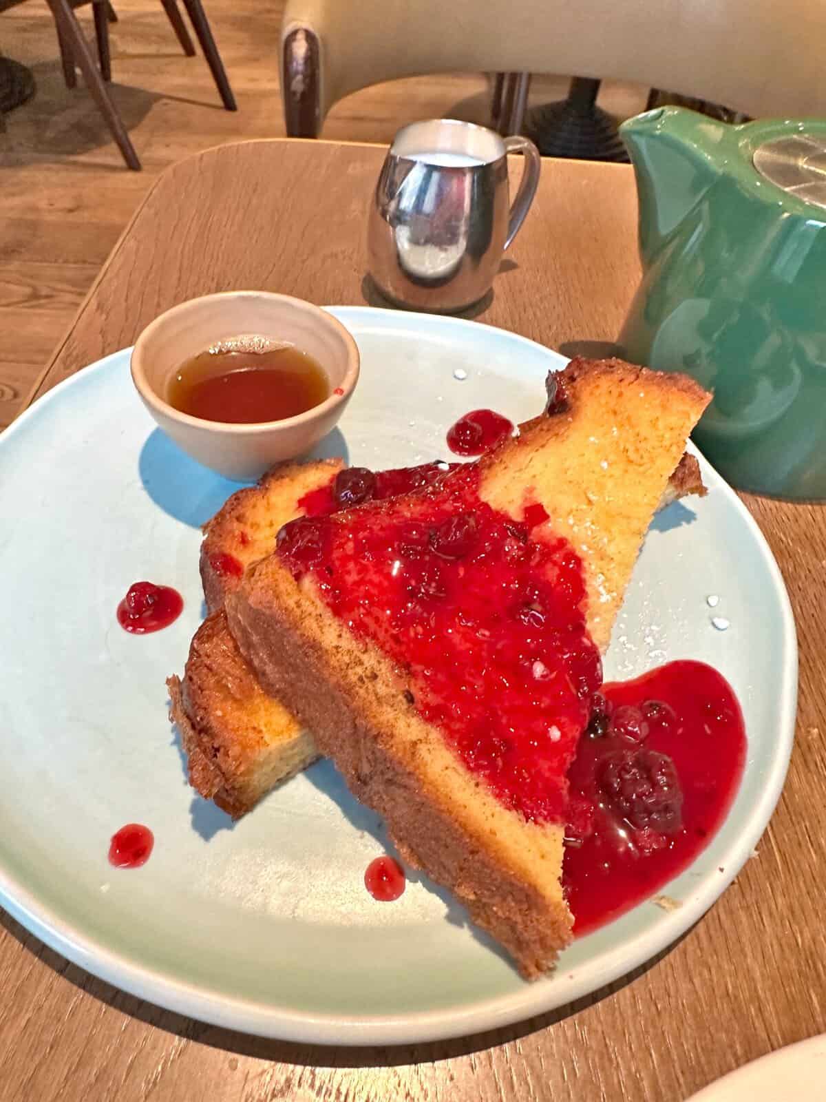 Two slices of French toast topped with berry compote at Andaz London Liverpool Street, served with a small bowl of syrup and a metal pitcher.