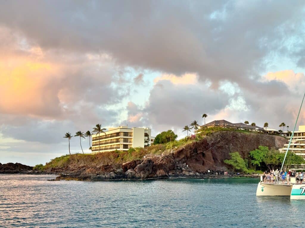 View of a rocky shoreline with a hotel, palm trees, and people on a boat in the foreground under a cloudy sky at sunset.