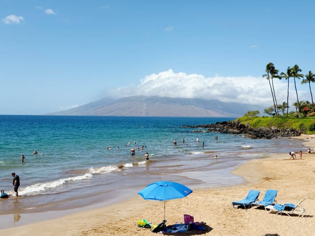 A sandy beach with people swimming in the blue ocean, two empty blue lounge chairs and an umbrella in the foreground, and a mountainous island in the background under a partly cloudy sky.
