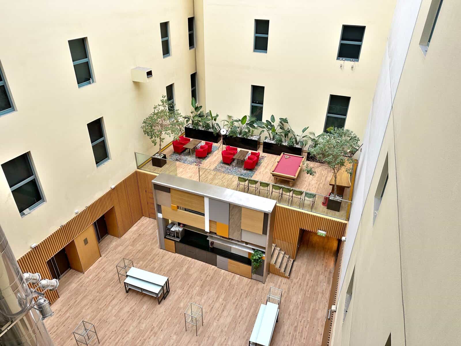 An elevated view of the Andaz London Liverpool Street atrium, featuring a lower-level lobby and an upper balcony with red seating, a pool table, and potted plants.
