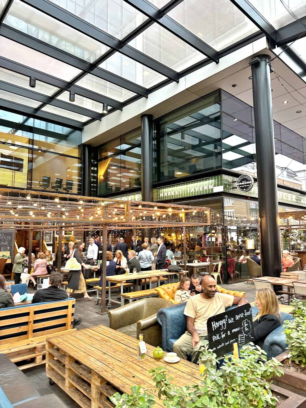 People sit at wooden pallet tables in an outdoor restaurant patio covered by a glass roof, a perfect spot during the best time to visit London's Old Spitalfield's Market