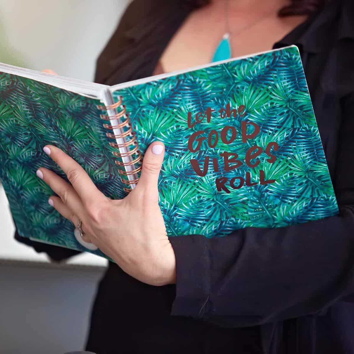 A person holds a notebook adorned with a green leaf pattern and the text "let the good vibes roll," perfect for jotting down a travel itinerary with room for spontaneity.
