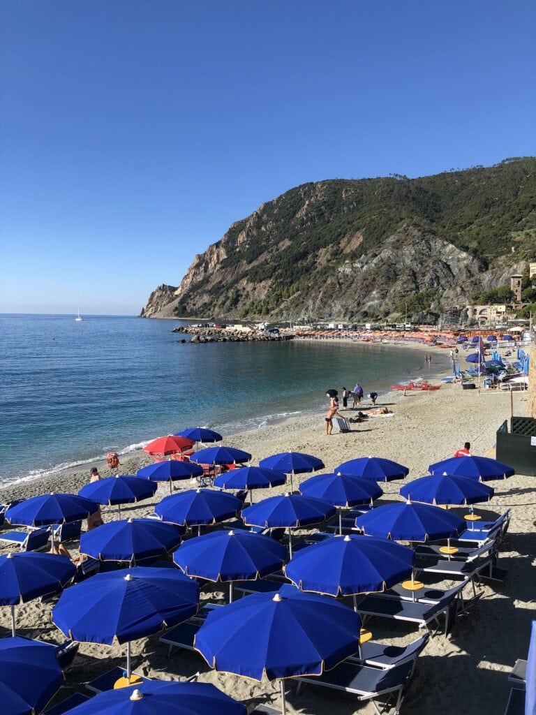 Rows of blue beach umbrellas and lounge chairs on a sandy beach, with people near the water and rocky cliffs in the background under a clear blue sky.