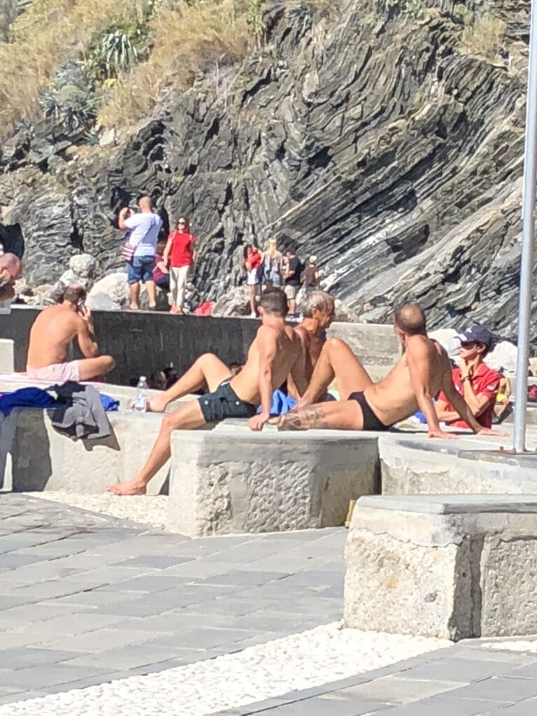 Several people are sunbathing and relaxing on stone benches near a rocky hillside, with others standing and walking in the background.