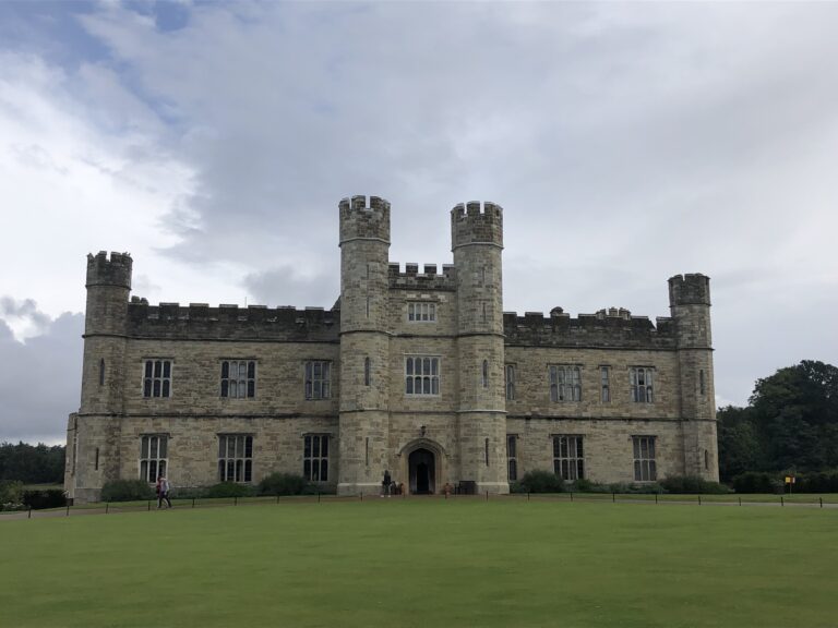 A large stone castle with four corner towers, arched entrance, and rectangular windows awaits travelers journeying from London to Leeds Castle, set on a manicured green lawn under a cloudy sky.
