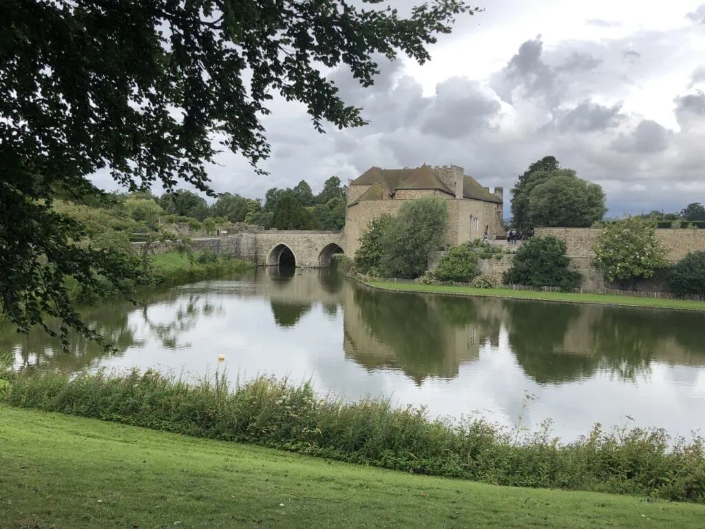 A stone bridge and castle sit by a calm river surrounded by greenery, reminiscent of the scenic route from London to Leeds Castle, with trees and cloudy sky reflected in the water.