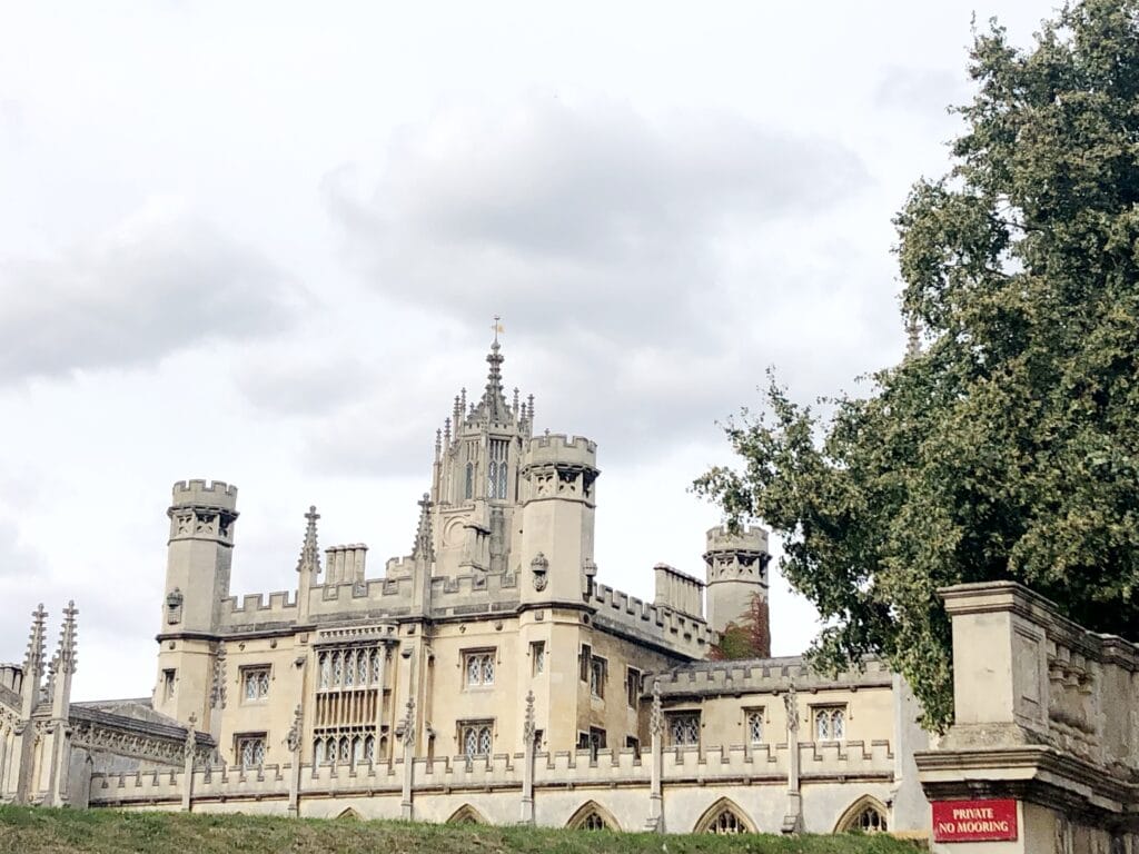 A large, historic Gothic-style building with towers and ornate windows stands behind a stone wall and gate under a cloudy sky—a stunning sight often enjoyed on a day trip from London to Cambridge.