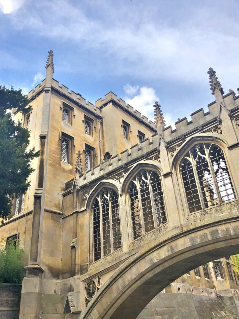 Stone building with arched windows and decorative window grilles, connected by a covered stone bridge—an iconic sight on a day trip from London to Cambridge, set against a partly cloudy sky.