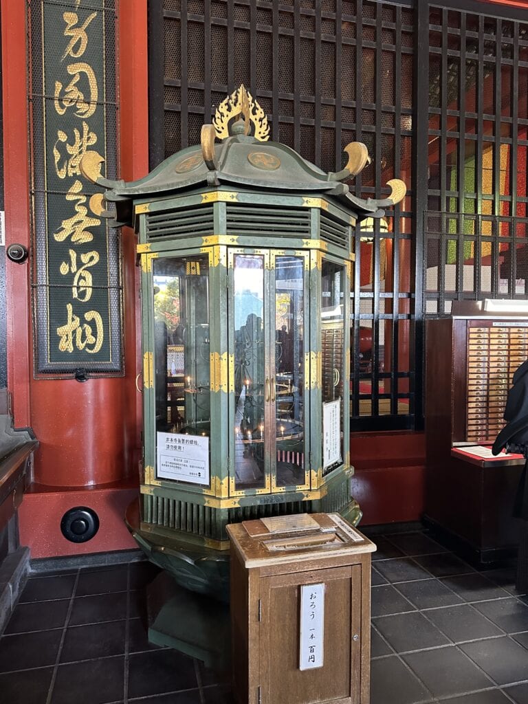 A glass and metal lantern stands indoors beside a wooden offering box and a decorative red and gold wall in a traditional Japanese temple, quietly embodying the traveling lessons found within sacred journeys.