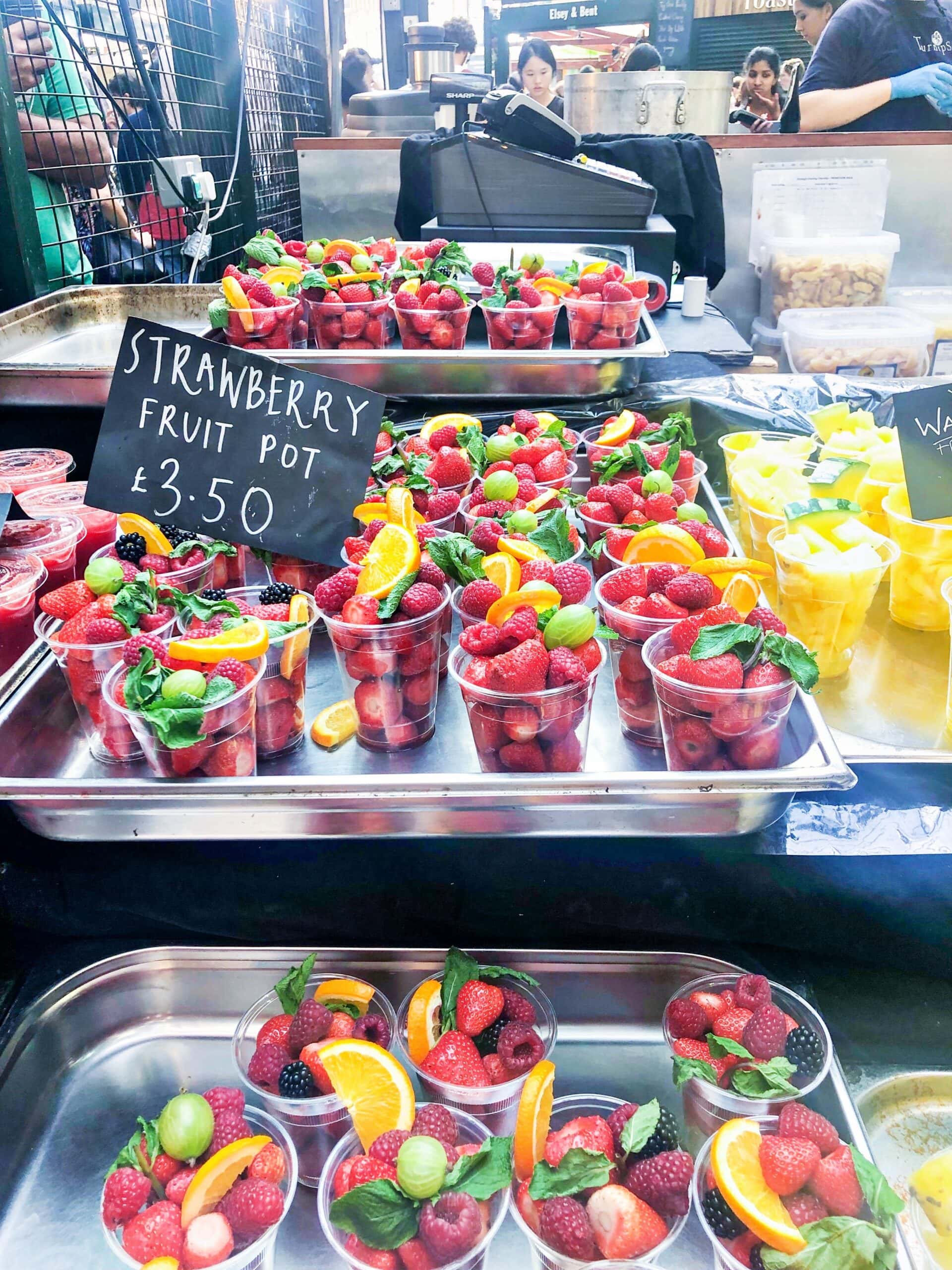 At one of the Best London Markets, clear plastic cups filled with strawberries, orange slices, and mint leaves are displayed on trays. A sign reads "Strawberry Fruit Pot £3.50.