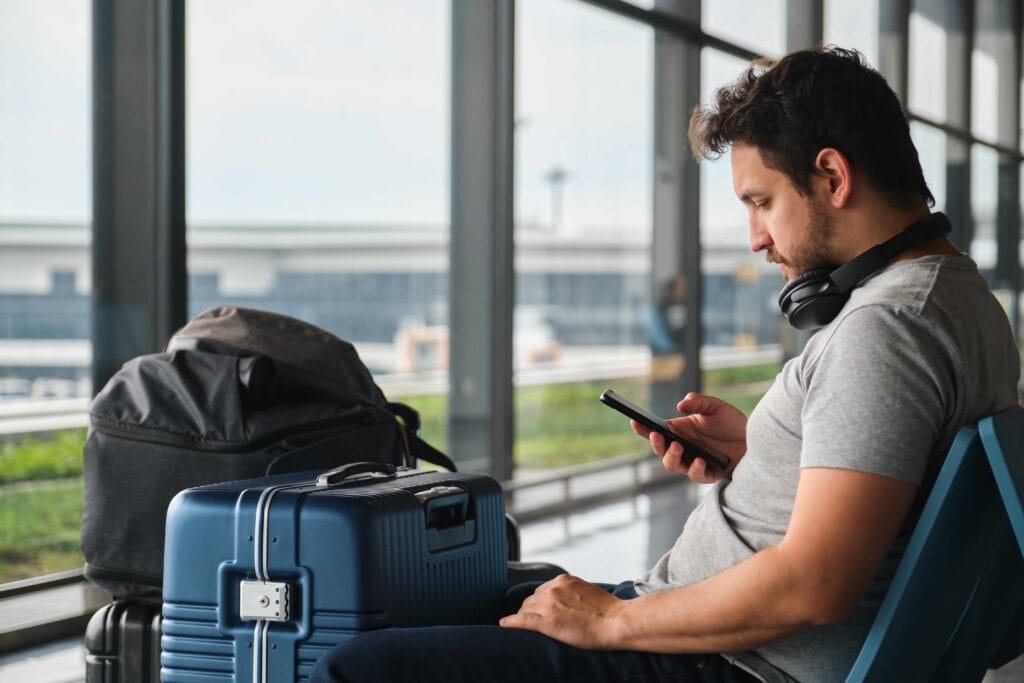 Man sitting in an airport terminal, looking at his phone with a suitcase, backpack, headphones around his neck, ready for his next adventure—perfectly prepared with an eSIM for European travel.