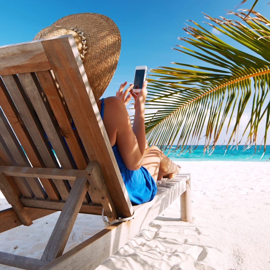 Person in a sunhat relaxing on a wooden lounge chair at a sandy beach, holding a smartphone with eSIM for European Travel, framed by palm leaves and the ocean in the background.