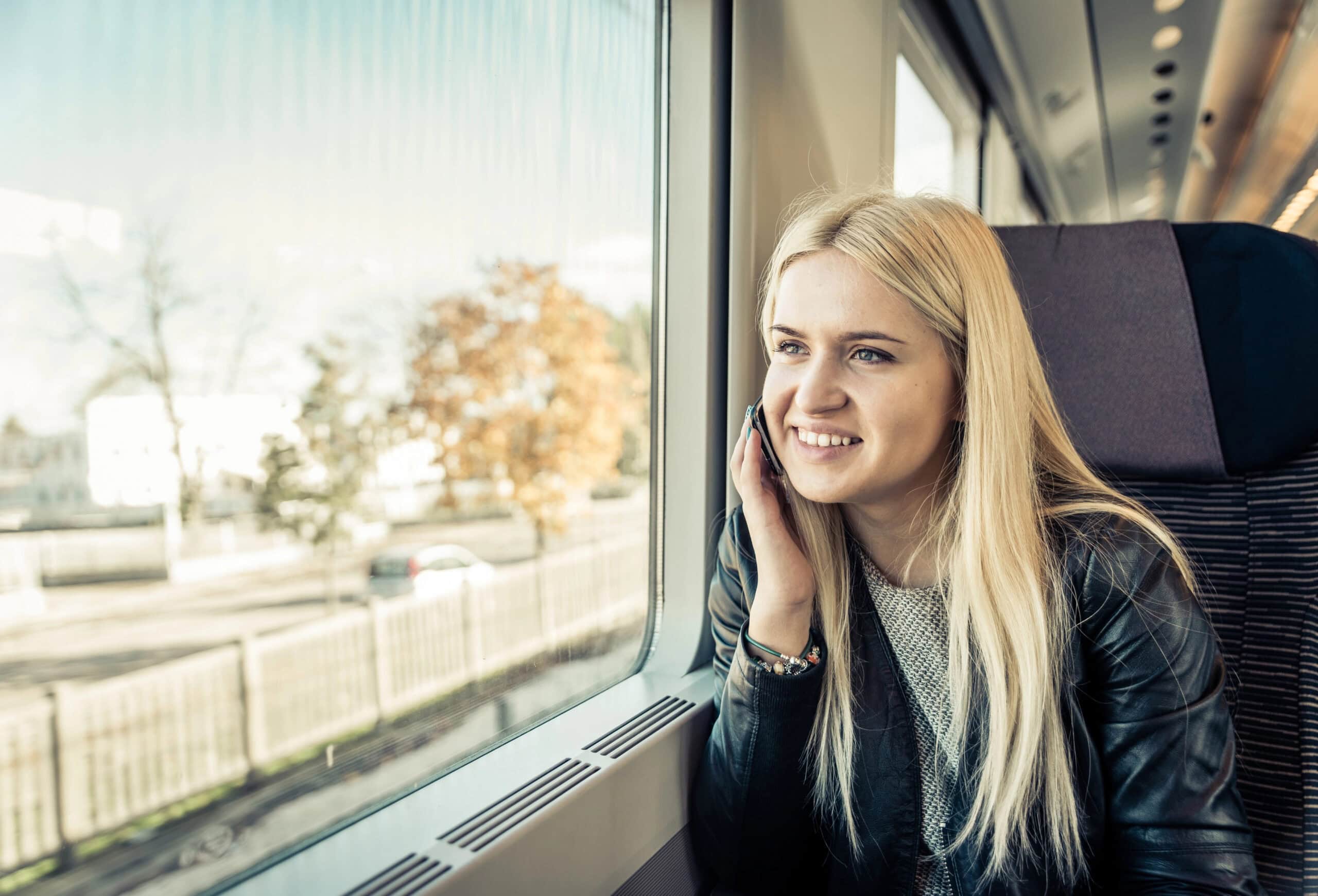 A woman with long blonde hair sits by a train window, smiling while talking on her phone using an eSIM for European travel. Trees and buildings pass by outside.