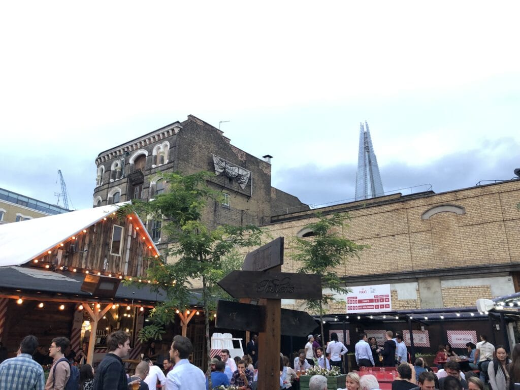 Outdoor urban scene with people socializing near a wooden cabin-style structure, brick buildings, and The Shard in the background—capturing the lively atmosphere often found at the best London markets under a cloudy sky.