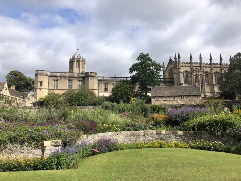 Historic stone building with a central tower and spires, surrounded by lush gardens and a manicured lawn under a partly cloudy sky.
