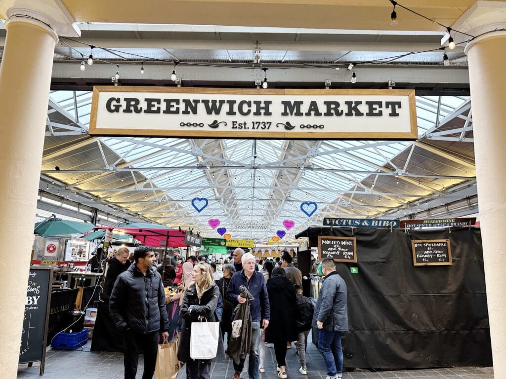 Entrance to Greenwich Market, one of the best London markets, with people walking and shopping at various stalls under a glass roof adorned with hanging lights and heart decorations.