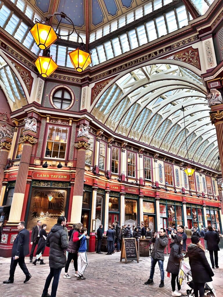 People stroll through one of the Best London Markets, an ornate, covered arcade with Victorian architecture, arched glass ceilings, and intricate details. Signs for "Lamb Tavern" adorn the charming storefronts.