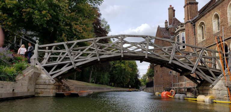 A wooden footbridge with a geometric design spans over a canal beside historic brick buildings in Cambridge, making it a scenic spot to visit on a day trip from London, with people near the water’s edge and kayaks floating below.