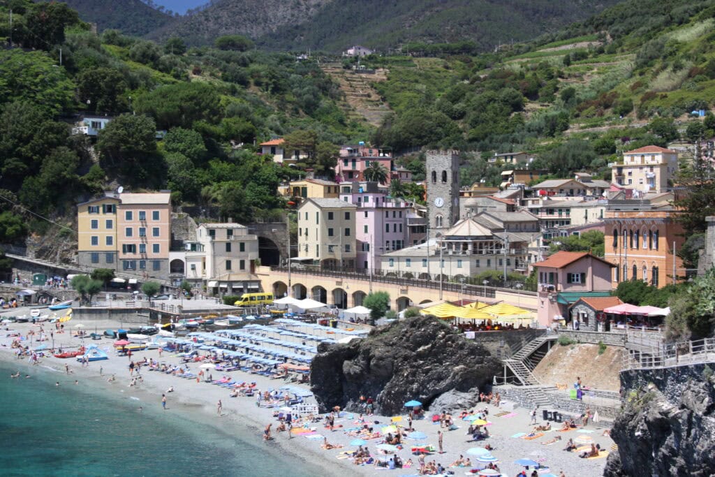 Crowded beach with umbrellas and sunbathers in front of pastel-colored buildings and green hills in a coastal village.