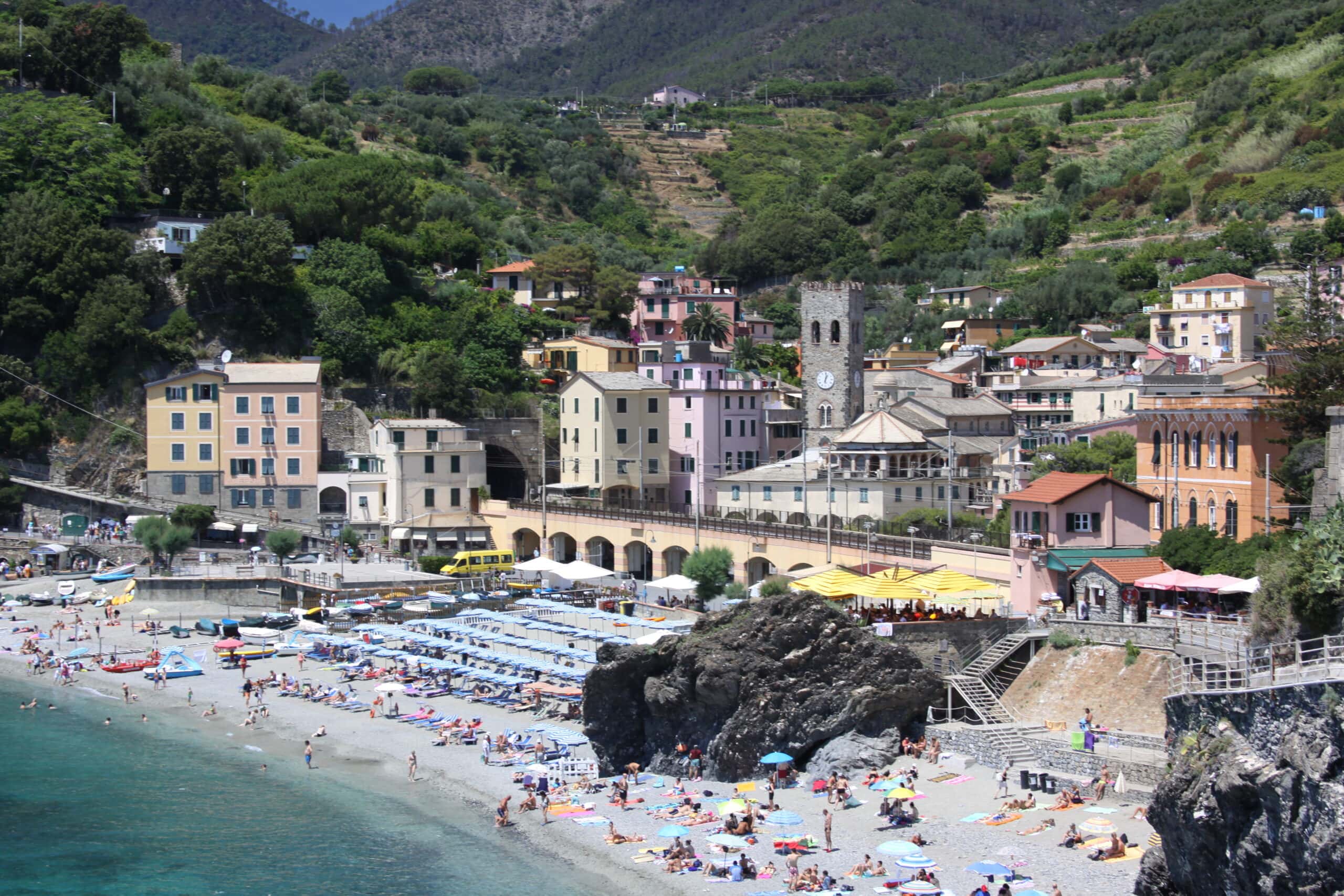 Crowded beach with umbrellas and sunbathers in front of pastel-colored buildings and green hills in a coastal village.