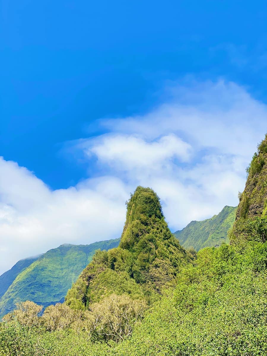 A green, cone-shaped hill surrounded by lush vegetation, with mountains and a blue sky scattered with clouds in the background—an iconic scene perfect for Maui for Adventurers seeking breathtaking landscapes.