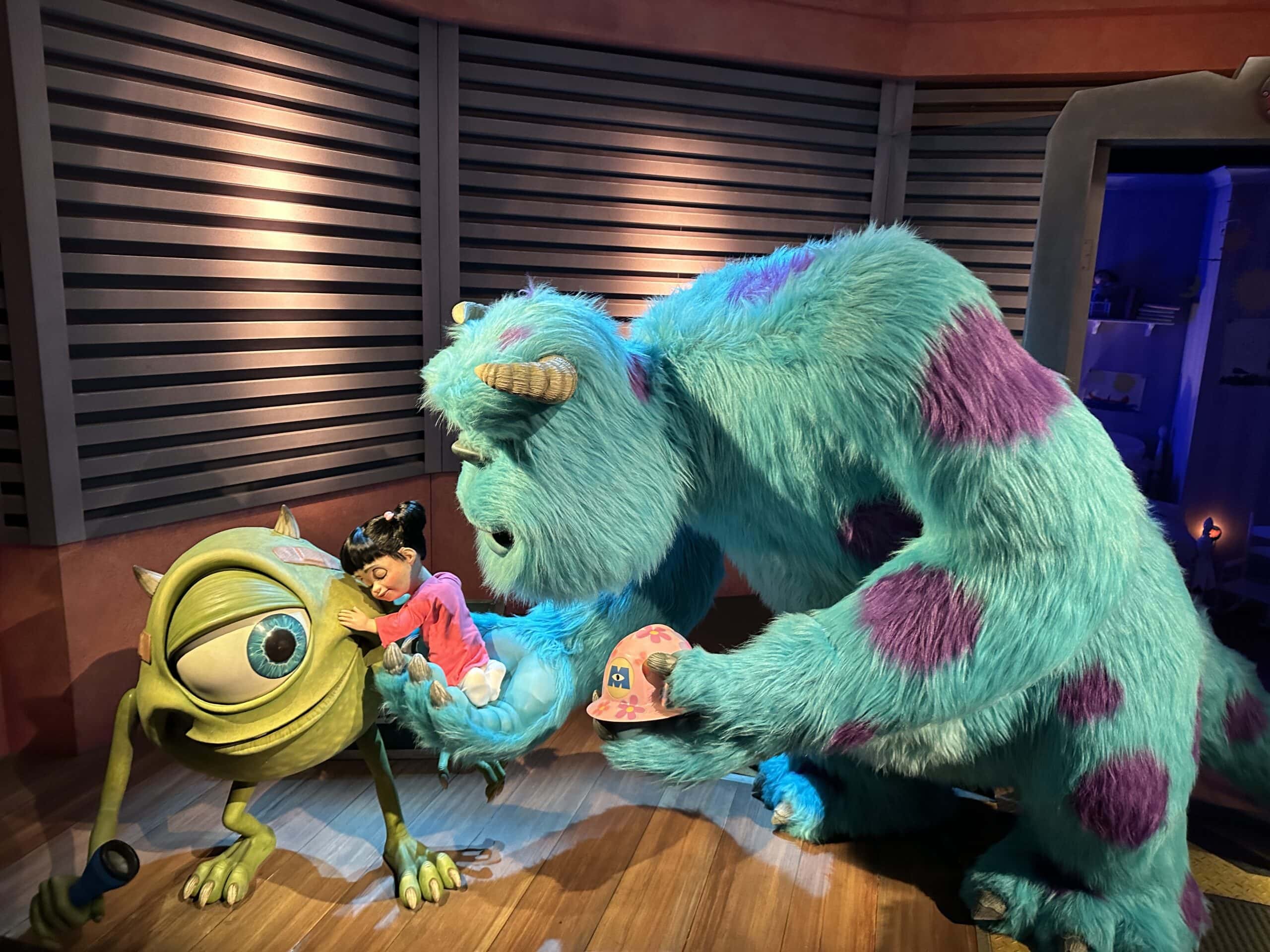 Two costumed characters, a green one-eyed monster and a large blue furry monster, interact with a small girl in a colorful room with striped walls.