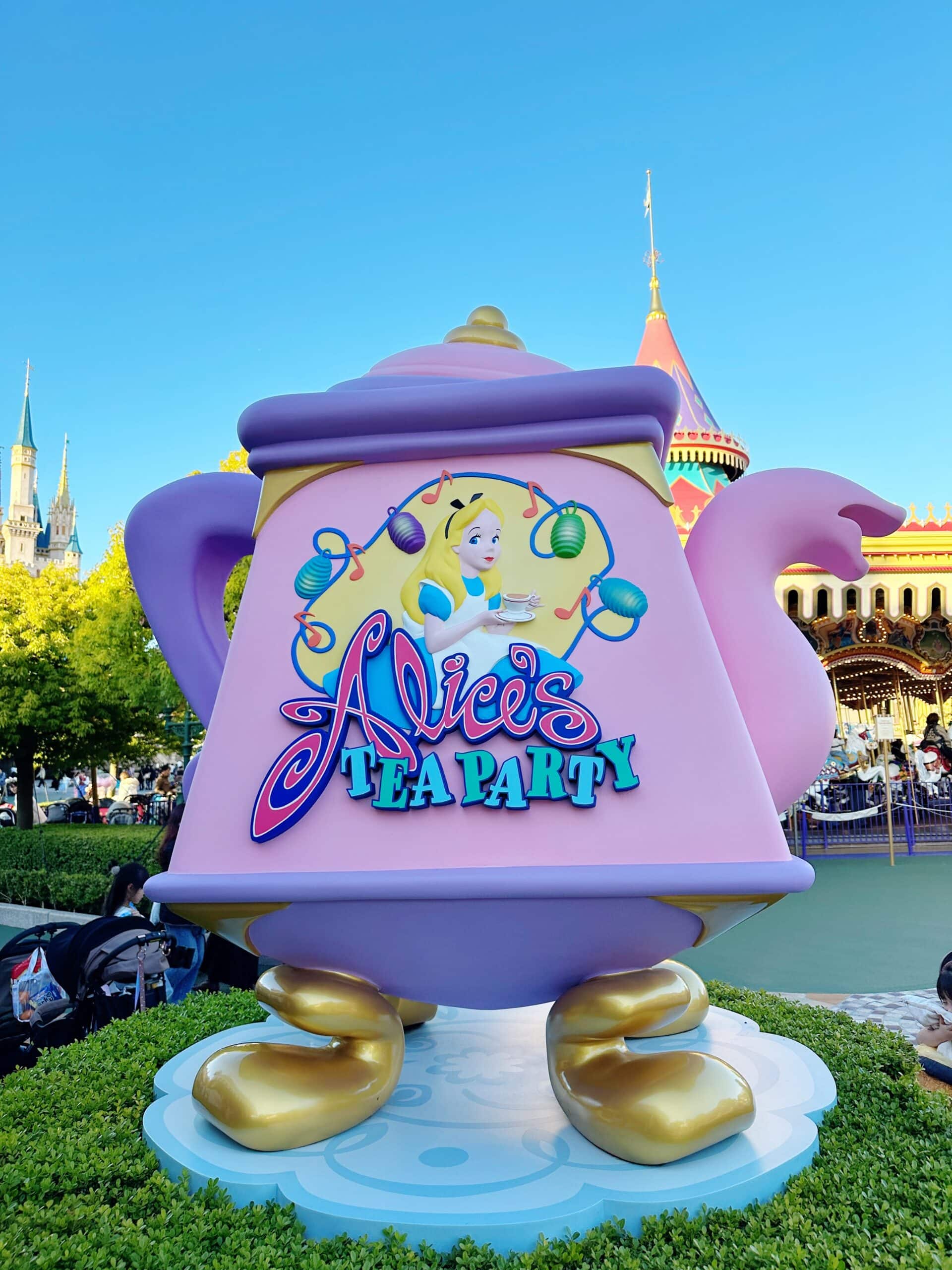 A large decorative teapot statue with "Alice's Tea Party" and an image of Alice, set outdoors with a castle and carousel in the background.