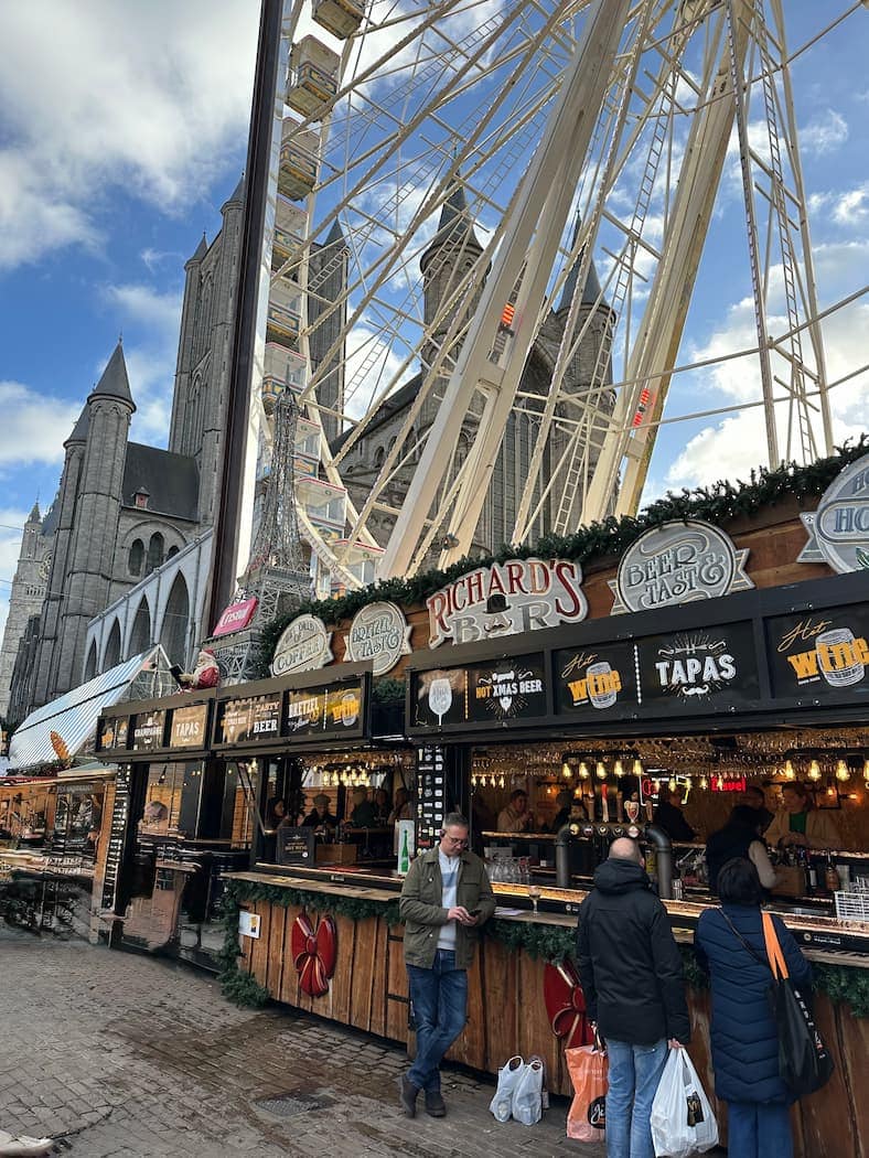 Outdoor market stalls selling food and drinks sit beneath a large Ferris wheel, with a church and cloudy blue sky in the background. People stand at the stalls.