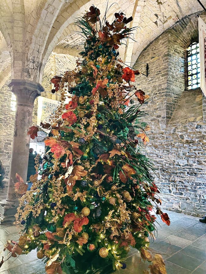 A decorated Christmas tree with gold and brown ornaments stands inside a stone-walled building with arched ceilings and a column.