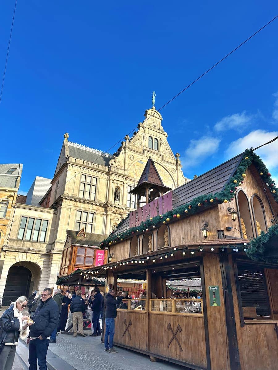 People gather near a wooden market stall decorated for the holidays in front of a historic building under a blue sky.