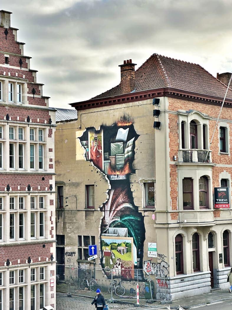 A mural on the side of a brick building depicts a scene of books, a sofa, and a waterfall, creating the illusion of an open wall in an urban street setting.