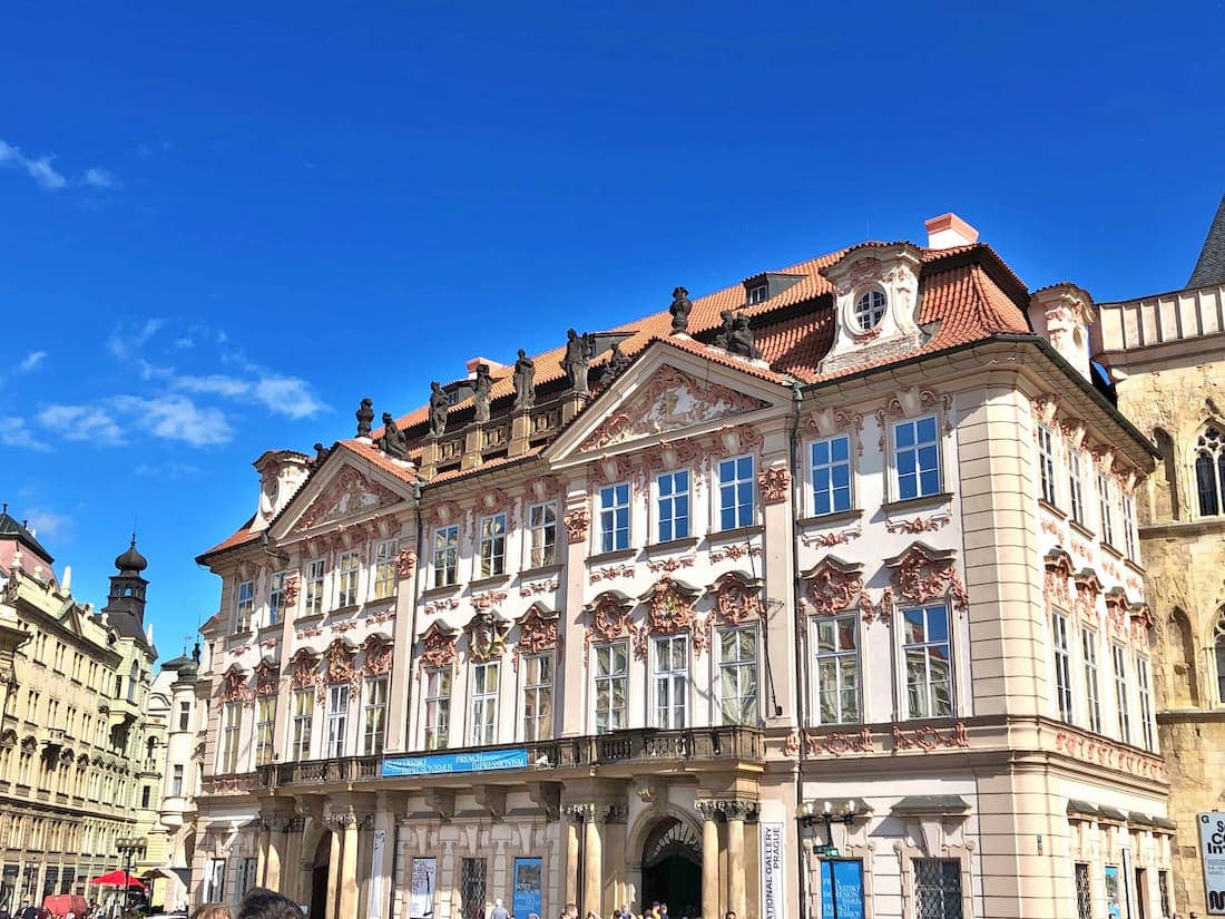 A large, ornate Baroque building with decorative facades and red-tiled roof stands under a clear blue sky, with people gathered in front.