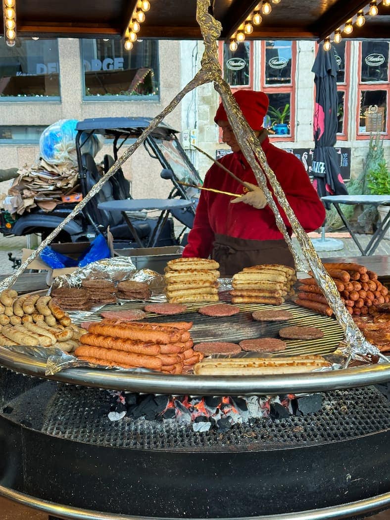 A person in a red hoodie grills various sausages, burgers, and meats on a large circular barbecue over an open flame at an outdoor market.