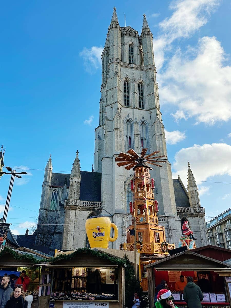 A tall Gothic-style church tower rises behind festive market stalls, a large mug decoration, and a wooden Christmas pyramid under a blue sky with scattered clouds.